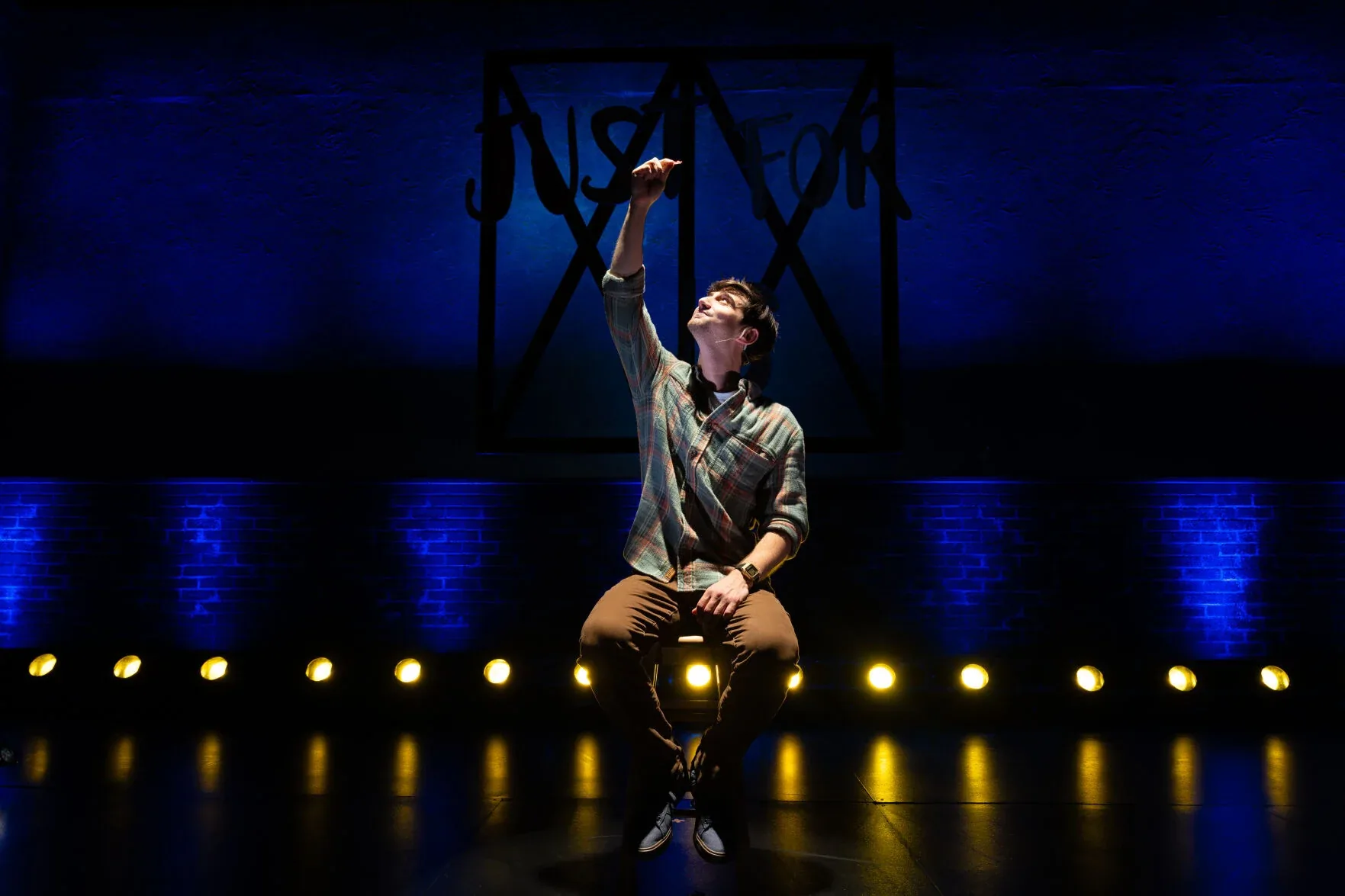 Jacob seated in a spotlight, reaching upward, in Just for Us